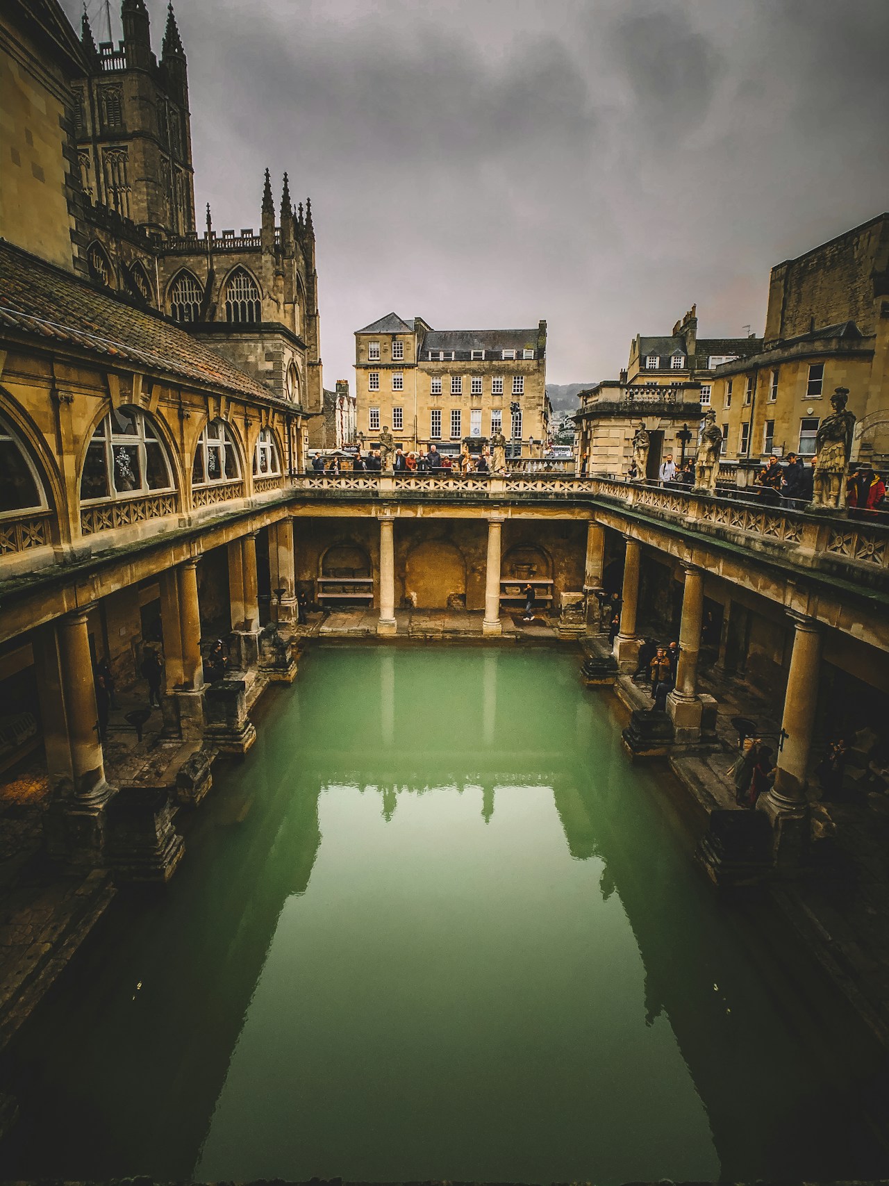 Green thermal pool at Roman Baths with stone columns and visitors.
